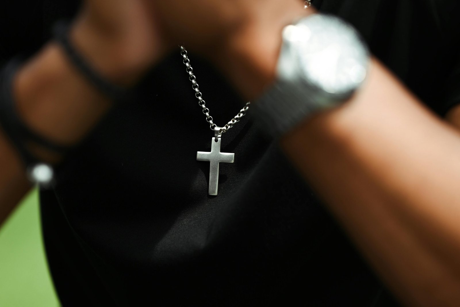 A close-up image of a silver cross pendant on a chain necklace worn by a person with a blurred watch in view.
